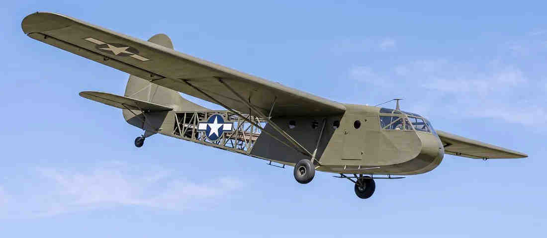 A clear, low-angle color photograph of a restored olive-drab Waco CG-4A glider soaring through a bright blue sky. The aircraft’s structural framework is partially visible through the fabric-covered fuselage, which prominently displays the white star and blue circle insignia. The Waco CG-4A glider A clear, low-angle color photograph of a restored olive-drab Waco CG-4A glider soaring through a bright blue sky. The aircraft’s structural framework is partially visible through the fabric-covered fuselage, which prominently displays the white star and blue circle insignia. The Waco CG-4A glider